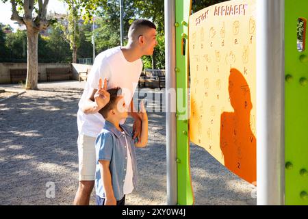 A young man with disabilities, including cerebral palsy and mild deafness, teaches sign language to a boy using an educational panel in the park Stock Photo