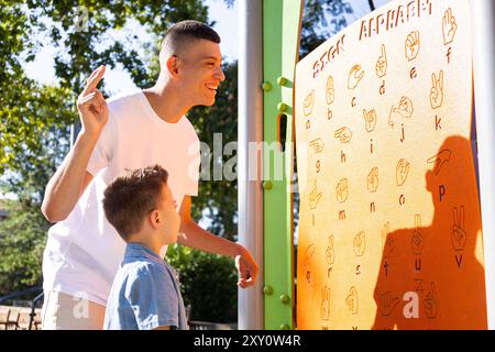 A young man with cerebral palsy and mild deafness joyfully teaches sign language to a young boy, using a colorful sign language chart outdoors Stock Photo