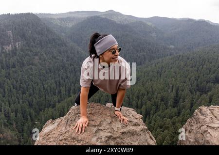 A smiling woman with sunglasses looks away while climbing a rock, showcasing a breathtaking forest view in the background. Stock Photo