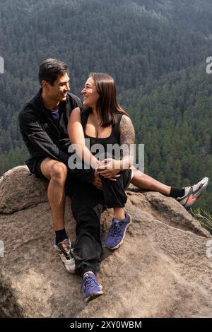 A male and female couple happily interacting on a rocky outcrop during a forest hike. They are looking at each other with smiles, surrounded by dense Stock Photo