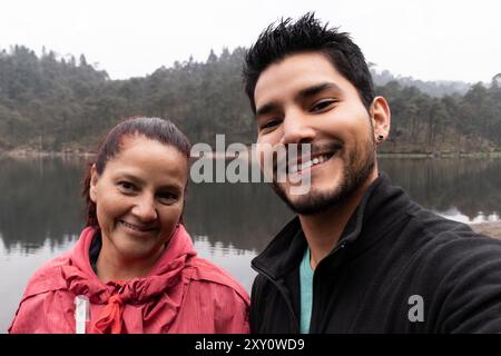 A middle-aged woman and a young man take a selfie against a serene lake backdrop on a misty, rainy day The quiet, green, foggy atmosphere gives a cool Stock Photo