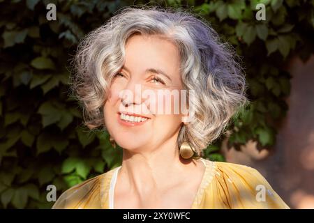 A joyful mature woman with curly gray hair smiles as she looking at camera, illuminated by sunlight. Her yellow dress and gold earrings complement the Stock Photo
