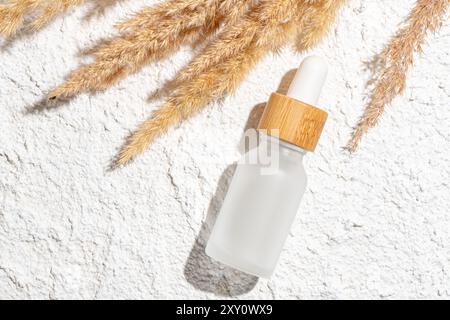 A glass skincare serum bottle with a wooden dropper cap rests on a textured stone background, surrounded by soft, golden pampas grass Stock Photo