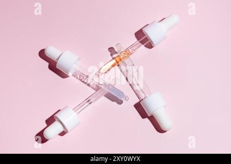 Top view of three transparent serum dropper bottles with white caps, two empty and one filled with amber liquid, artistically arranged in an X-shape o Stock Photo