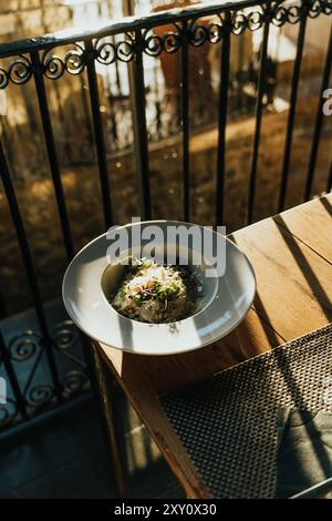 A gourmet dish beautifully presented on a white plate, positioned on a wooden table against a wrought iron balcony railing, bathed in warm sunlight Stock Photo
