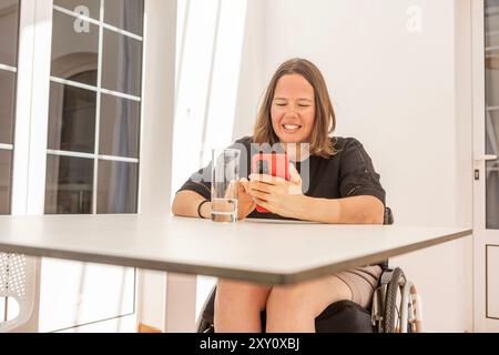 A cheerful woman in a wheelchair engages with her smartphone at a modern office, reflecting a theme of active wheelchair users handling business Stock Photo