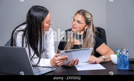Two Latin Hispanic female lawyers sit at a desk engaged in discussion, with one holding a digital tablet and papers, in a professional office setting. Stock Photo