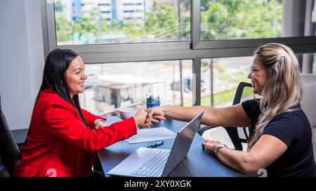 Two professional Latin Hispanic women, one in a red blazer and another in a black shirt, are seen shaking hands over a business deal in a modern offic Stock Photo