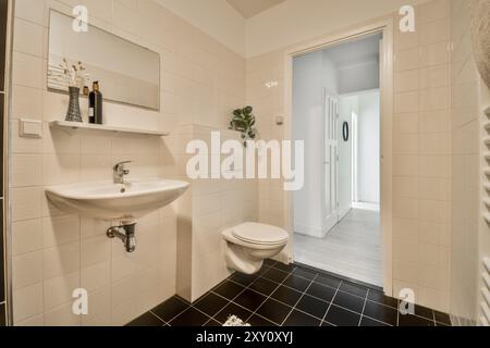 A sleek modern bathroom featuring white tiled walls with contrasting black floor tiles, a pedestal sink, toilet, and decorative shelving. Stock Photo