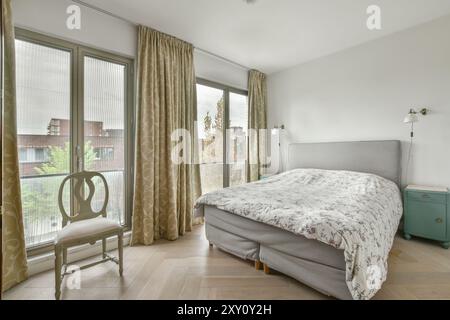 A modern bedroom with large windows draped in patterned curtains, showcasing a comfortable bed, decorative chair, and clean, minimalist design. Stock Photo