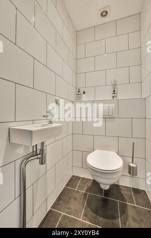 Modern white tiled bathroom interior with a stylish washbasin and toilet on dark tiled flooring. Stock Photo