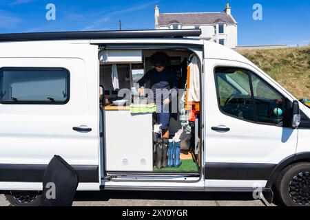 A young man enjoys the convenience of his camper van by preparing lunch inside the compact, well-equipped kitchen on a sunny summer day, surrounded by Stock Photo