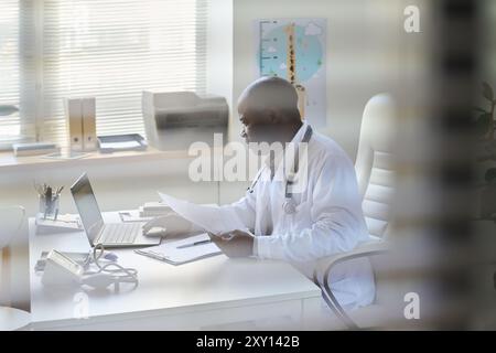 Medical professional analyzing documents in an office environment with bright sunlight coming through window blinds. Surroundings include various offi Stock Photo