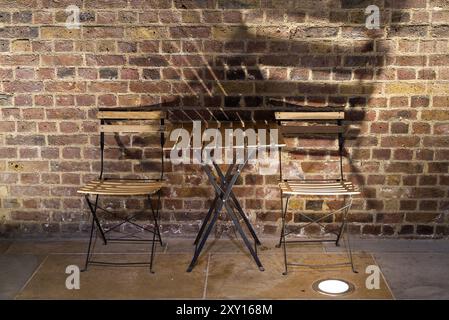 A wooden table and chairs of a cafe against a brick wall with lights creating shadows Stock Photo