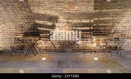 Two wooden table and chairs of a cafe against a brick wall with lights creating shadows Stock Photo