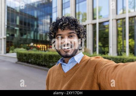 Happy young man taking selfie with smartphone, smiling broadly in front of modern glass office building. Casual attire with orange sweater and blue shirt reflects relaxed urban style, expressing joy Stock Photo