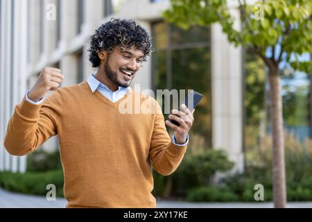 Smiling young man in casual attire celebrating success with raised fist and smartphone in hand. Energetic expression shows happiness, achievement, and positivity. Stock Photo