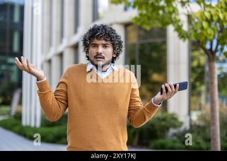 Young man with curly hair shrugs while holding smartphone, expressing confusion or uncertainty. Stock Photo