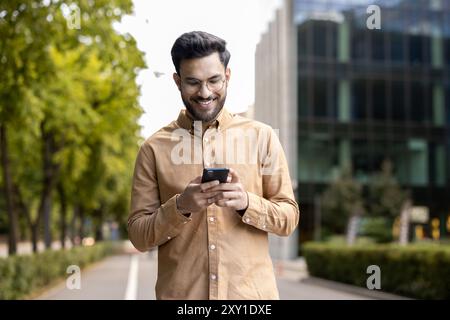 Smiling man wearing glasses and casual clothes using smartphone outdoors. Enjoying technology in urban setting with modern buildings. Capturing moment connectivity and communication through phone. Stock Photo