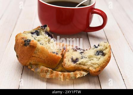 Closeup of a blueberry muffin with a cup of coffee in the background. The muffin is broken in half on a rustic white wood table. Stock Photo