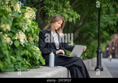 Happy satisfied woman freelancer with laptop working online outside in park sitting near bush plants Stock Photo