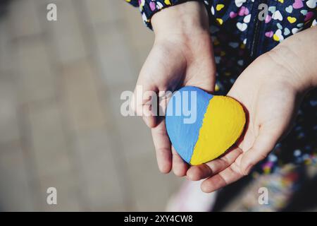 Small little kid hold heart shaped stone with Ukraine blue-yellow national flag or anthem. Girl love native country. Child stand for Ukrainians. Stop Stock Photo