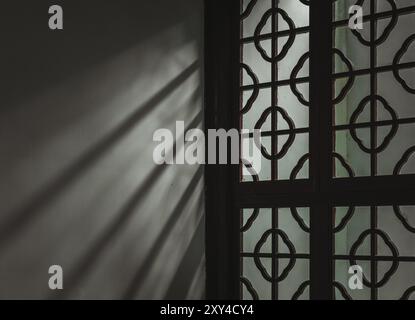 Sunlight streams through a window with a delicate lattice design, casting elongated shadows on the wall. Stock Photo