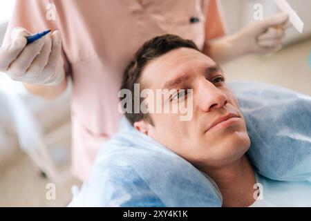 Closeup face of handsome male patient with hair loss problem lying on couch, nurse marking with pencil new hairline on head before hair Stock Photo