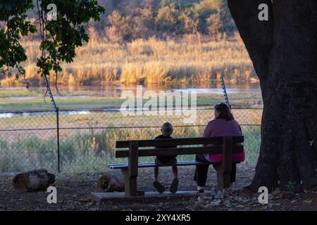 Lower-sabie, South Africa - an unidentified mother and son watching over the river from a bench inside the rest camp in the Kruger Park Stock Photo