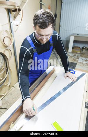 A bearded tired handsome carpenter on a table with a pencil draws a sign on the board. At the workplace there is an ordinary master dressed in blue ca Stock Photo