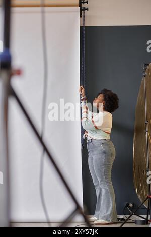 Minimal full length portrait of Black female photographer setting up backgrounds in photo studio preparing for creative project Stock Photo