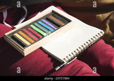 A close-up on the girl's legs is a notebook with a pencil and a set of pastel crayons for drawing. The concept of creativity and drawing Stock Photo