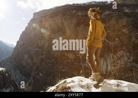 Full-length portrait A girl photographer in sunglasses and a big fur hat and a yellow knitted sweater stands against the background of high rocks in t Stock Photo