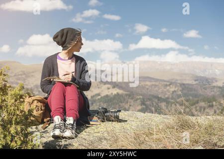 Stylish girl designer painter in the mountains sitting on a rock draws in a notepad at sunset Stock Photo