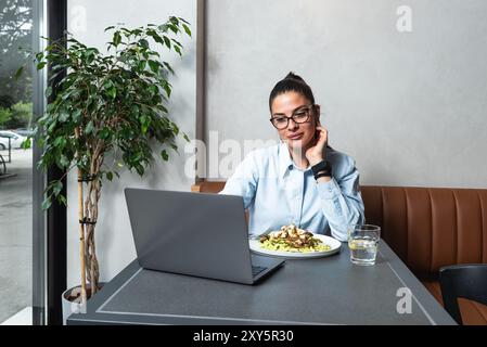 Young business woman working remotely from cafeteria during lunch break. Female office worker using laptop computer while she eating at restaurant to Stock Photo