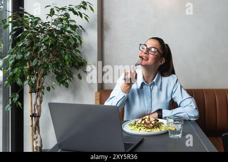 Young business woman working remotely from cafeteria during lunch break. Female office worker using laptop computer while she eating at restaurant to Stock Photo