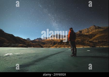 A brooding man stands on the surface of a frozen lake illuminated by moonlight high in the mountains against the backdrop of epic rocks. The concept o Stock Photo