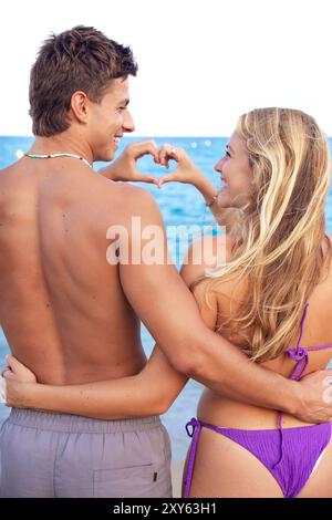 Young couple making heart shape with their hands on the beach looking at camera Stock Photo
