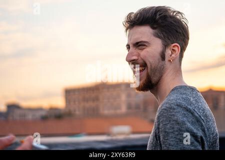 Candid portrait of a cheerful young bearded man laughing heartily on a city rooftop at sunset. Natural, genuine emotion captured in warm evening light Stock Photo
