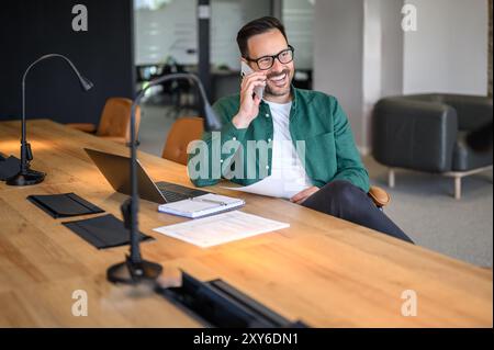 Happy adult businessman discussing over phone call and looking away while sitting at desk in office Stock Photo