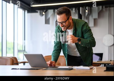 Smiling male freelancer drinking coffee and checking e-mails over wireless computer on desk in office Stock Photo