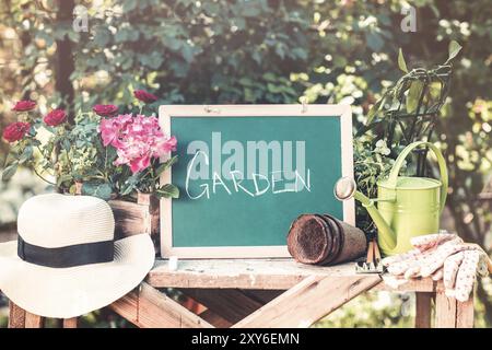 Small chalkboard with inscriptions Garden on wooden table among green plants in clay pots straw hat and metal watering can Stock Photo