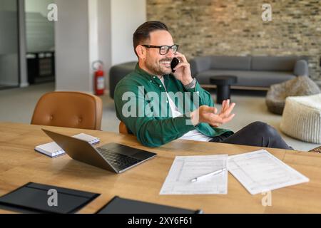 Male programmer smiling and discussing project over phone call while working on laptop at desk in office Stock Photo