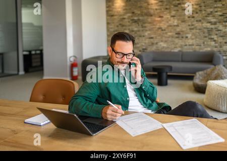 Male project manager in glasses talking over mobile phone and writing notes on paper at his workplace Stock Photo