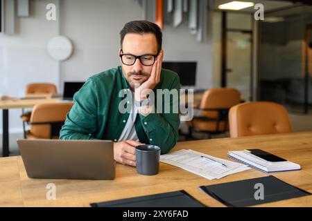 Serious young businessman with coffee cup looking at laptop computer while working at desk in office Stock Photo