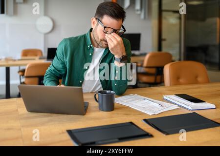 Exhausted male manager rubbing eyes in stress while working over project on laptop at his workplace Stock Photo