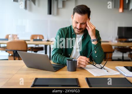 Young businessman holding eyeglasses and touching head in pain while working over laptop in office Stock Photo