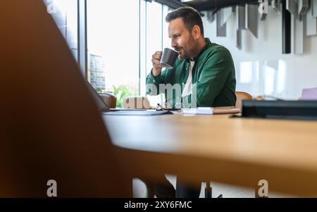 Male project manager drinking coffee while relaxing at wooden desk in modern office during breaks Stock Photo