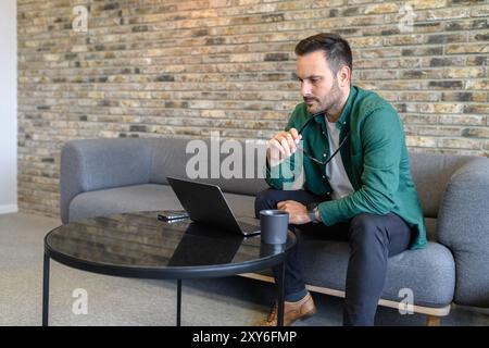 Pensive male professional holding eyeglasses and looking at laptop while sitting on sofa in office Stock Photo