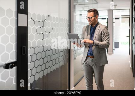 Smiling young businessman in eyeglasses working on his computer while walking in office corridor Stock Photo